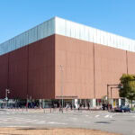 Visiting Yokohama Cup Noodles Museum A large, modern, cubic brick building with ECQUI MUSEUM written at the entrance stands at a city intersection—reminiscent of the iconic Cup Noodles Museum—with pedestrians, cars, and trees nearby on a clear day.