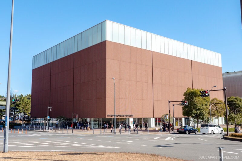 A large, modern, cubic brick building with ECQUI MUSEUM written at the entrance stands at a city intersection&mdash;reminiscent of the iconic Cup Noodles Museum&mdash;with pedestrians, cars, and trees nearby on a clear day.