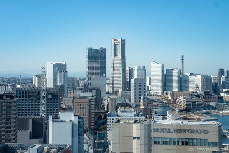 A cityscape view of Yokohama, Japan, featuring modern high-rise buildings under a clear blue sky. The prominent Hotel New Grand and the iconic Yokohama Marine Tower are visible in the foreground.