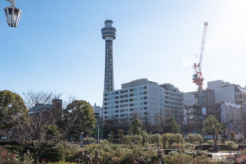 Yokohama Marine Tower rises above modern buildings and a construction crane, with a park filled with trees and flowers in the foreground under a clear blue sky.