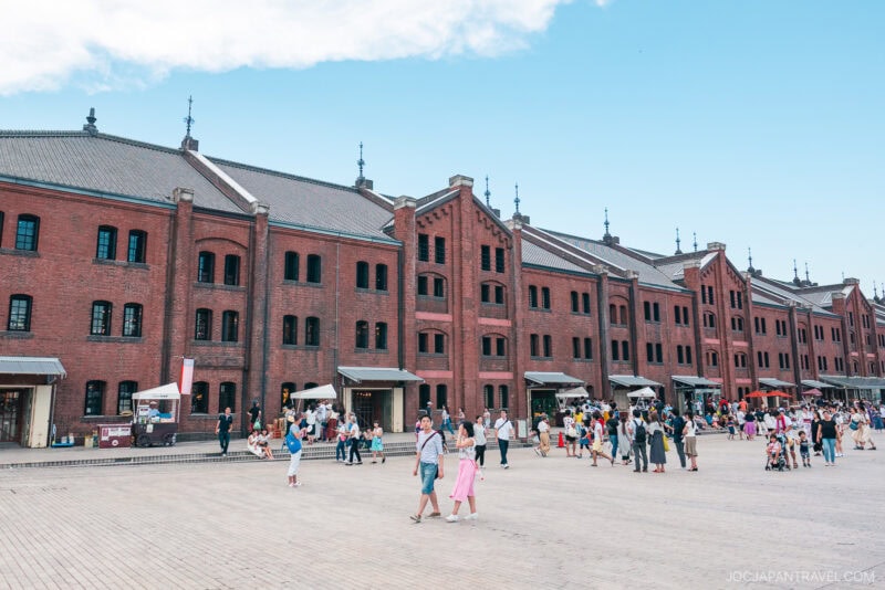 The Yokohama Red Brick Warehouse is a large red brick building with multiple peaked roofs and tall windows, surrounded by many people walking and gathering in a spacious open plaza under a partly cloudy blue sky.