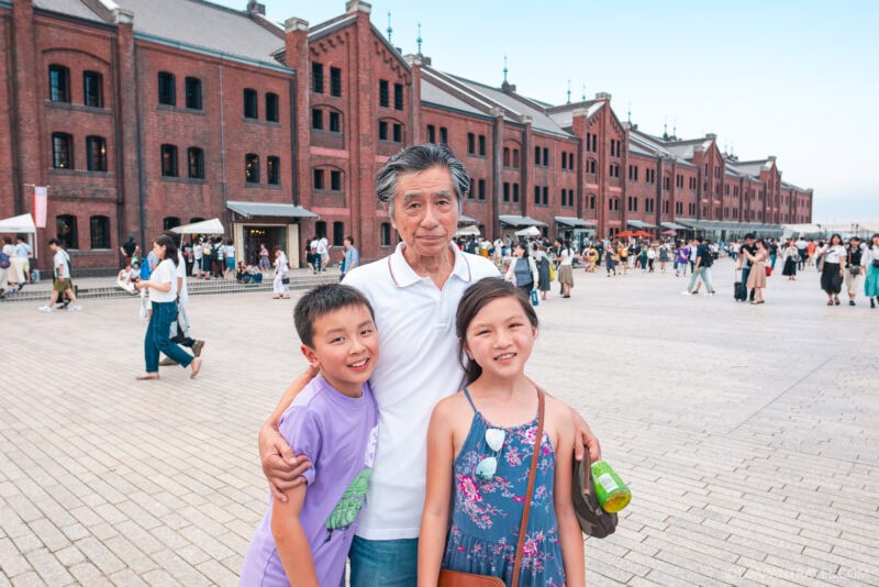 An older man stands outdoors with his arms around a smiling boy and girl in front of the Yokohama Red Brick Warehouse, as crowds of people walk and gather in the background.
