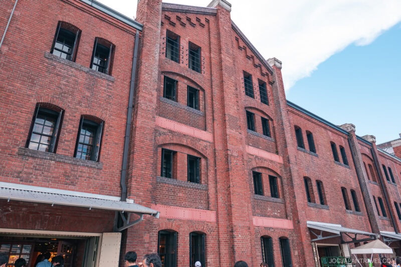 A large red brick building with black-framed windows, arched details, and people gathered near the entrance on a sunny day with a partly cloudy sky, resembling the iconic Yokohama Red Brick Warehouse.