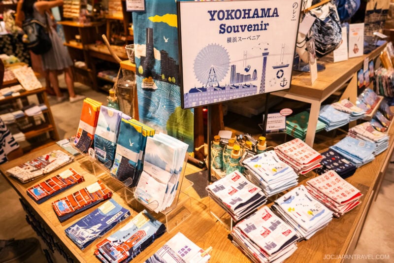 A display of Yokohama-themed souvenirs, including folded towels and cloths with various prints, arranged on wooden shelves in a store at Yokohama Red Brick Warehouse. A sign above highlights Yokohama Souvenir with city illustrations.