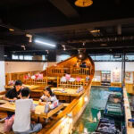 People dine at wooden tables inside Zauo, a bright and modern restaurant featuring a unique boat-shaped seating area and fish tanks beside the tables, with hanging lights and visible menu signs enhancing the lively atmosphere.