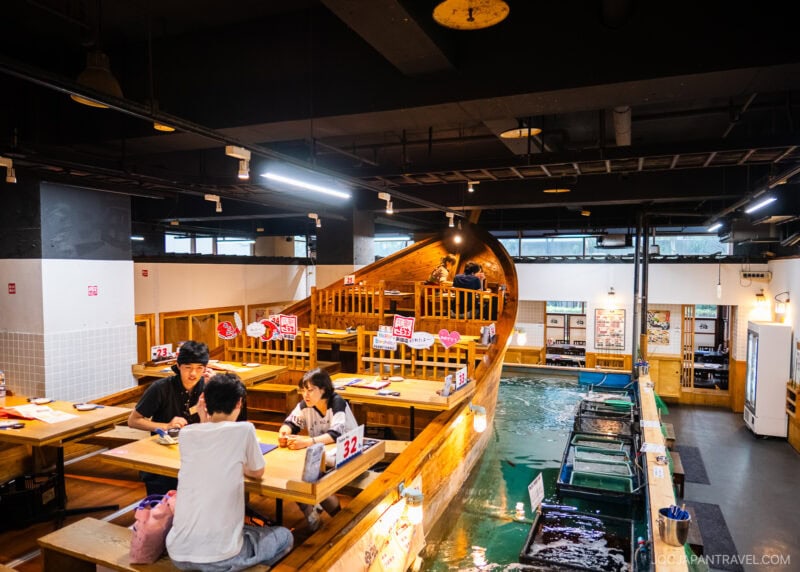People dine at wooden tables inside Zauo, a bright and modern restaurant featuring a unique boat-shaped seating area and fish tanks beside the tables, with hanging lights and visible menu signs enhancing the lively atmosphere.