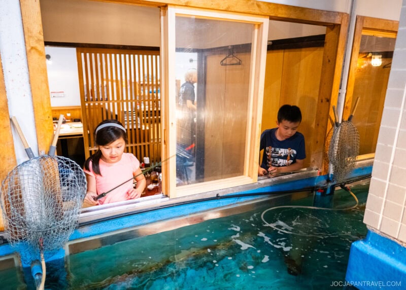 Two children stand by a large indoor fish tank at Zauo, holding fishing rods and peering into the water. Behind them, a wooden partition and window separate the fishing area from the dining space.