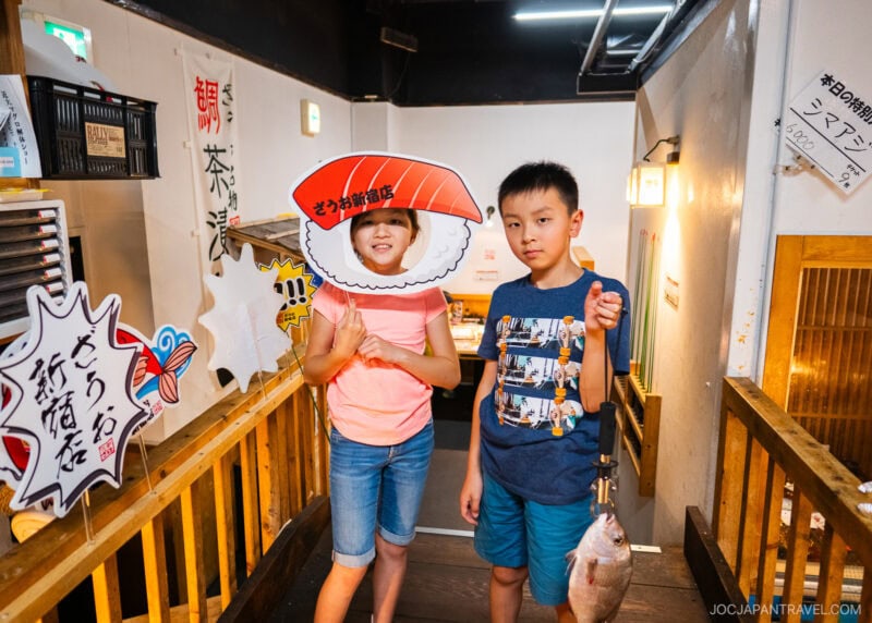 Two children stand indoors at Zauo; one poses with a sushi cutout and a peace sign, while the other holds a fish and looks at the camera. Japanese signs decorate the background.