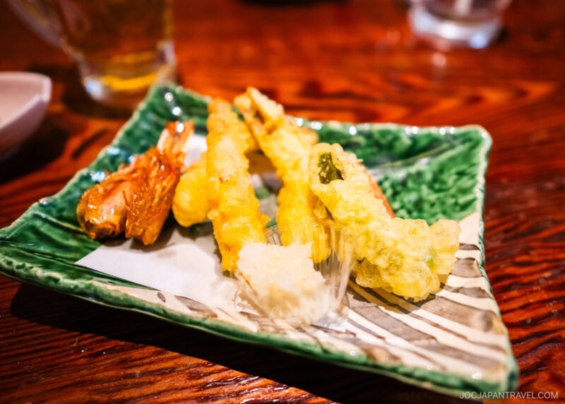 A green ceramic plate at Zauo holds assorted tempura, including shrimp and vegetables, with a small portion of grated daikon on the side. The plate rests on a wooden table.