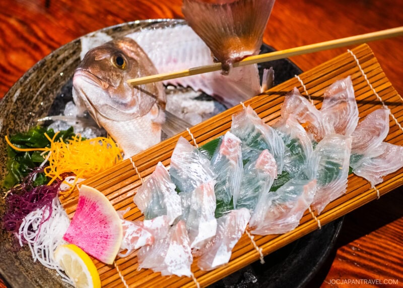 A plate of sashimi at Zauo features thinly sliced raw fish arranged on a bamboo mat, garnished with vegetables. In the background, a whole fish head and tail are presented on a bed of ice for an authentic dining experience.