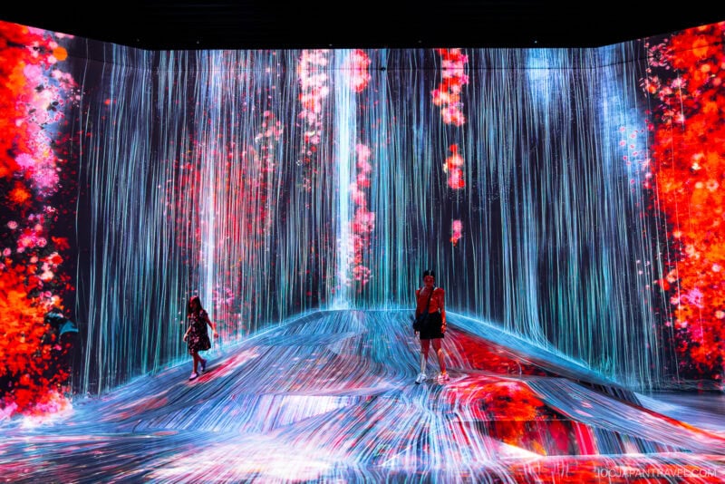 Two people stand amid vibrant digital art at teamLab Borderless, with cascading blue lines resembling a waterfall and bright orange and red abstract shapes surrounding them on the walls and floor.