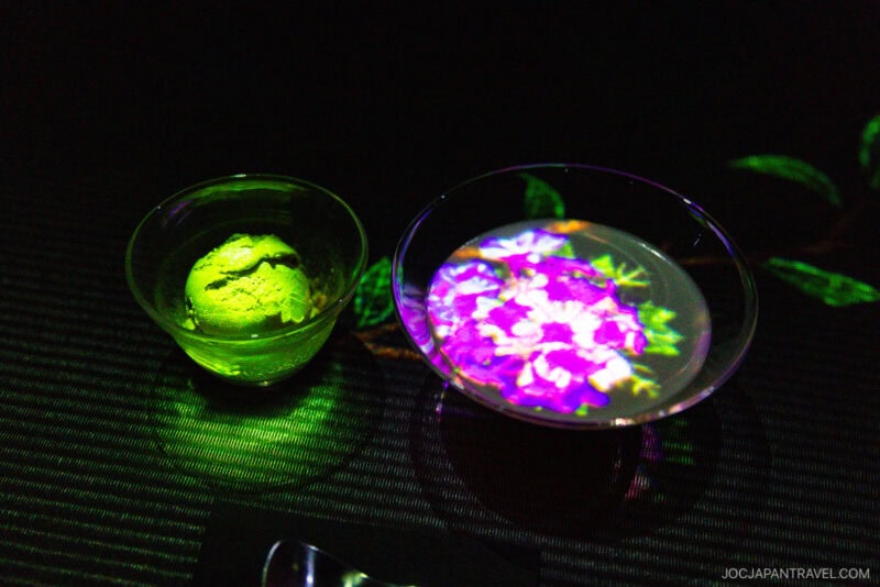 A glass bowl of green ice cream sits beside a glass dish of water with colorful, illuminated floral projections—reminiscent of teamLab Borderless—set against a dark background.