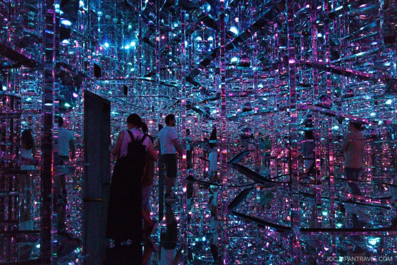 People walk through a mirrored room at teamLab Borderless, filled with colorful, glowing lights in purple, blue, and pink, creating infinite reflections and a surreal, immersive environment.