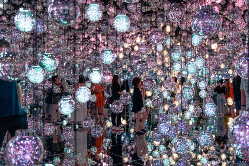 People walk through an art installation at teamLab Borderless, filled with hundreds of hanging, glowing, multicolored glass orbs. The mirrored floor and walls create infinite reflections, enhancing the immersive, sparkling atmosphere.