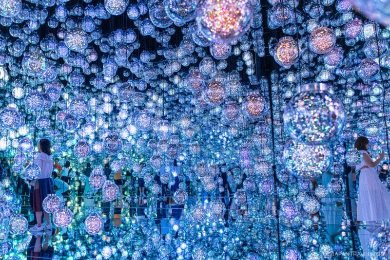 People stand in a mirrored room at teamLab Borderless, surrounded by countless glowing, multicolored orbs that create a mesmerizing, immersive light display with infinite reflections in every direction.
