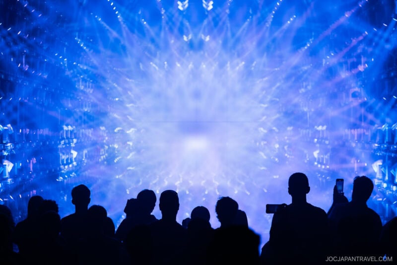 Silhouette of a crowd facing a vibrant blue light display with radiating patterns, creating a dynamic, immersive visual effect reminiscent of teamLab Borderless. Some people are capturing the scene with their phones.