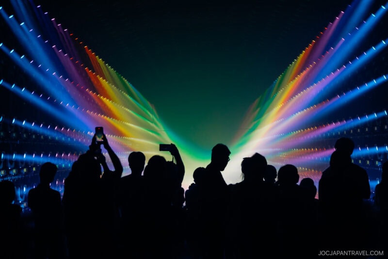 Silhouetted crowd stands before a vivid rainbow light display forming a wing-like shape at teamLab Borderless, with some people holding up phones to capture the scene. The atmosphere is vibrant and immersive.