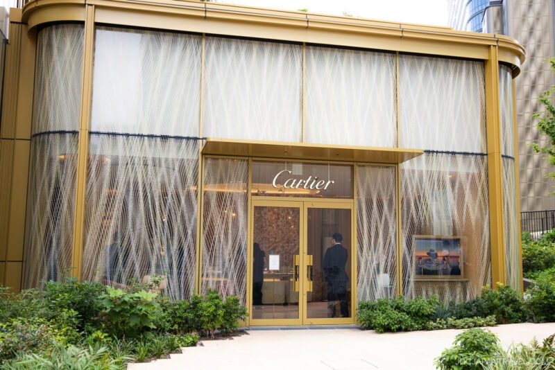 A modern Cartier storefront with gold accents, large glass doors, and decorative vertical patterns inspired by teamLab Borderless, surrounded by lush greenery and landscaping.