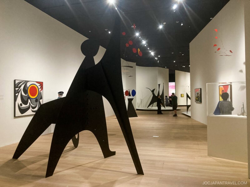 A modern art gallery, inspired by teamLab Borderless, features abstract sculptures, colorful paintings, and mobiles. Visitors observe the artwork under bright lights, while a security guard stands nearby on wooden floors and amid white walls.
