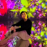 teamLab Planets Tokyo (チームラボプラネッツ東京) – An Immersive Art Experience A young person sits barefoot on a reflective floor at teamLab Planets Tokyo, surrounded by vivid, colorful digital art projections of flowers and leaves, creating a mesmerizing, immersive effect.