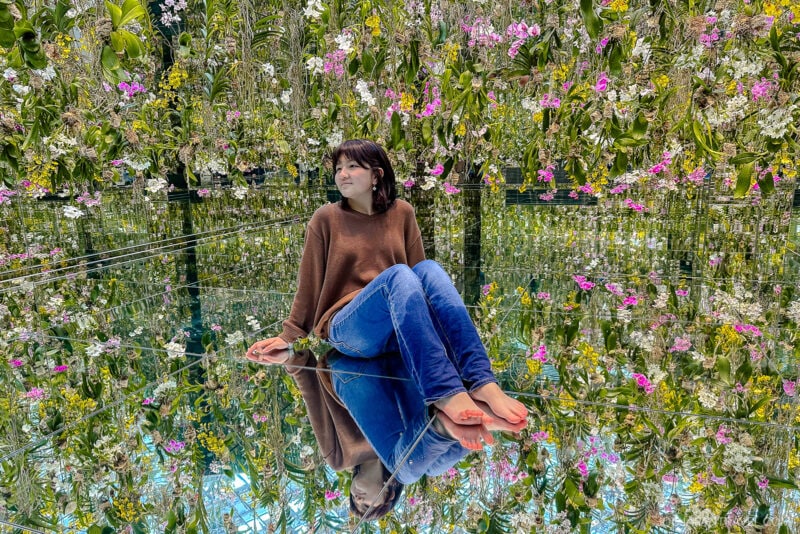 A woman sits barefoot on a reflective floor at teamLab Planets Tokyo, surrounded by hanging flowers and green plants, creating a mirrored, immersive garden effect. She wears a brown sweater and blue jeans, appearing calm and contemplative.