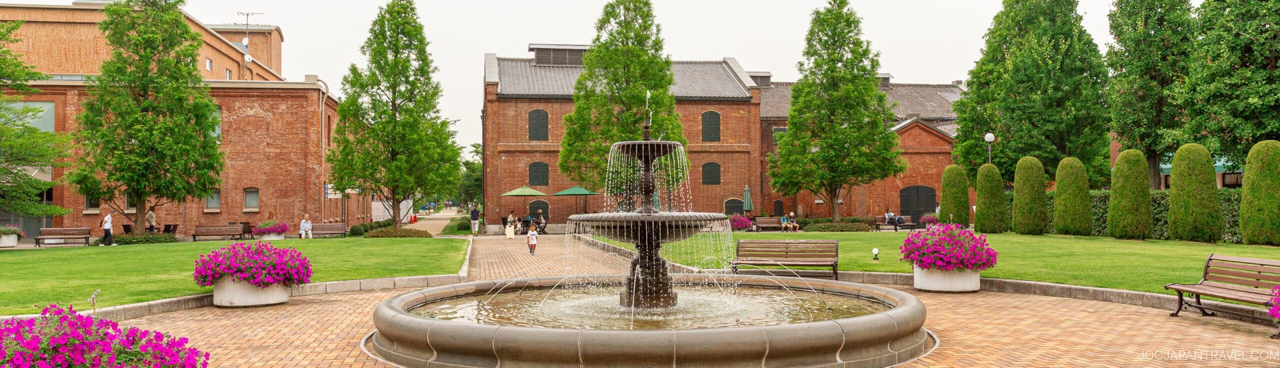 A round fountain surrounded by benches and purple flower pots stands in the center of a brick courtyard in Aichi prefecture, bordered by green lawns, tall trees, and red brick buildings in the background.