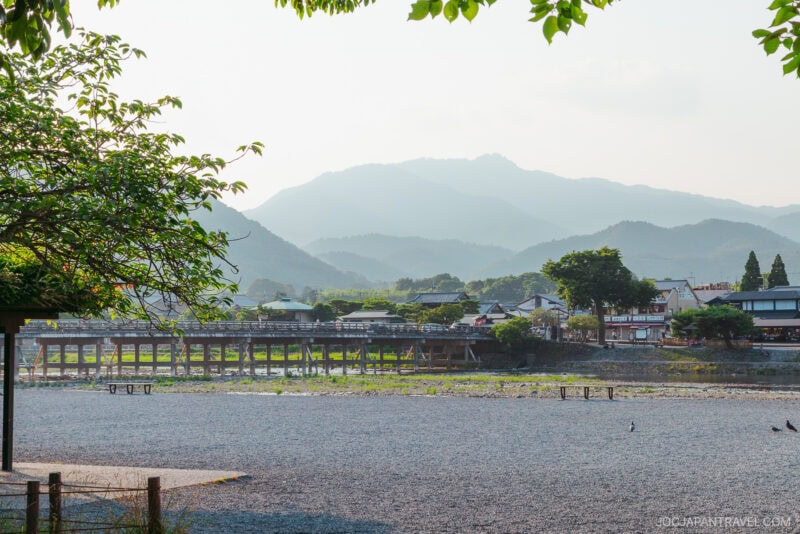 A scenic view of a river with a wide pebble shore, the iconic Togetsukyo Bridge, and houses nestled among trees, set against green mountains under a hazy sky with leafy branches framing the top.