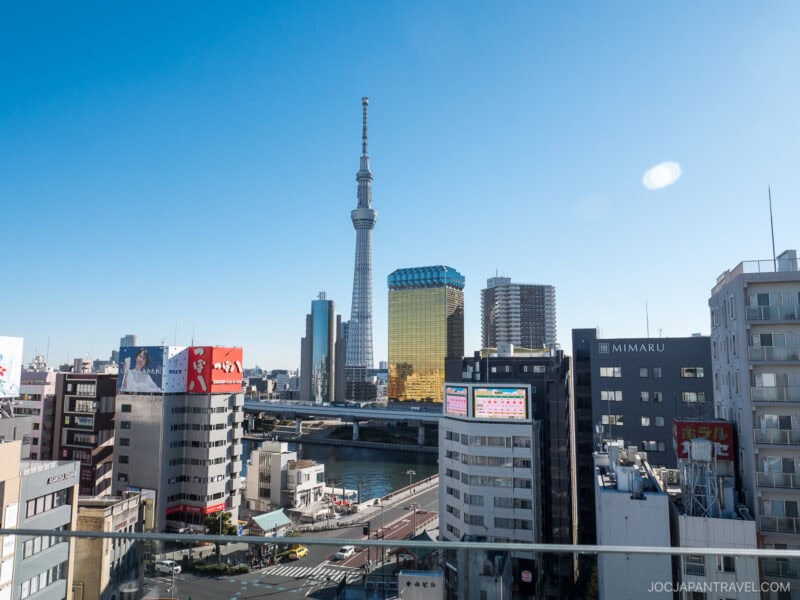 View of Tokyo cityscape with the Tokyo Skytree tower centered, surrounded by modern buildings, a river, and clear blue sky in the background.