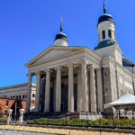 One Perfect Day in Baltimore: History, Harbor Views & Maryland Blue Crab A large, historic Baltimore church with tall columns, twin domes, and a classical façade stands under a clear blue sky. A white tent is set up nearby, with surrounding buildings visible in the background.