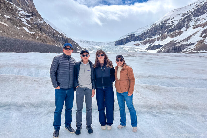 Four people wearing winter jackets and sunglasses stand close together, smiling on a snowy glacier in Banff National Park, with mountains and a partly cloudy sky in the background.