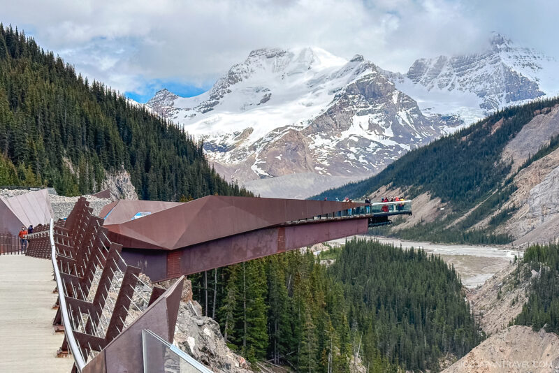 A group of people stands on a glass-floored observation platform extending over a forested valley in Banff National Park, with snow-capped mountains and a cloudy sky in the background.