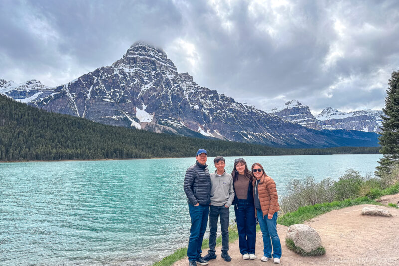Four people stand smiling by a turquoise lake in Banff National Park, with snow-capped mountains and a cloudy sky in the background, surrounded by trees and a rocky shoreline.