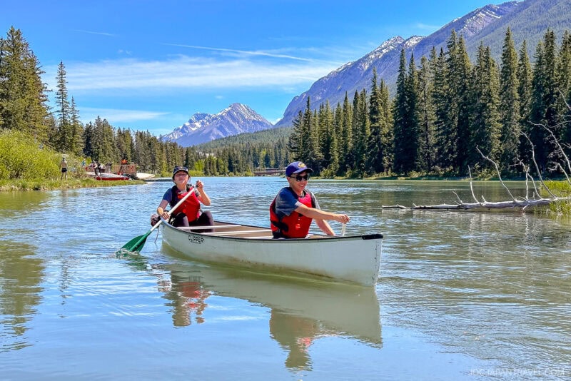 Two people in life jackets paddle a canoe on a calm river in Banff National Park, surrounded by pine trees and mountains under a blue sky. Another group is visible in the background on the water.