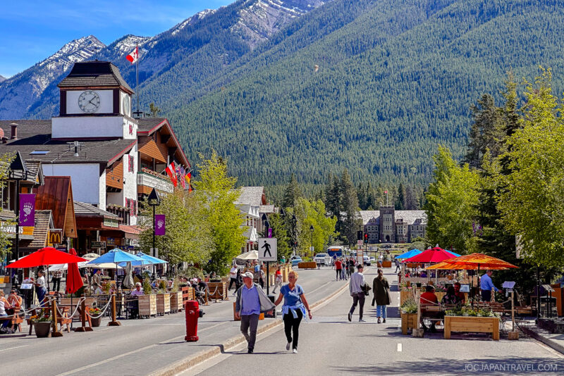 Pedestrians walk along a lively street lined with shops, outdoor seating, and colorful umbrellas in a mountain town near Banff National Park, with snow-capped peaks and pine trees towering in the background.