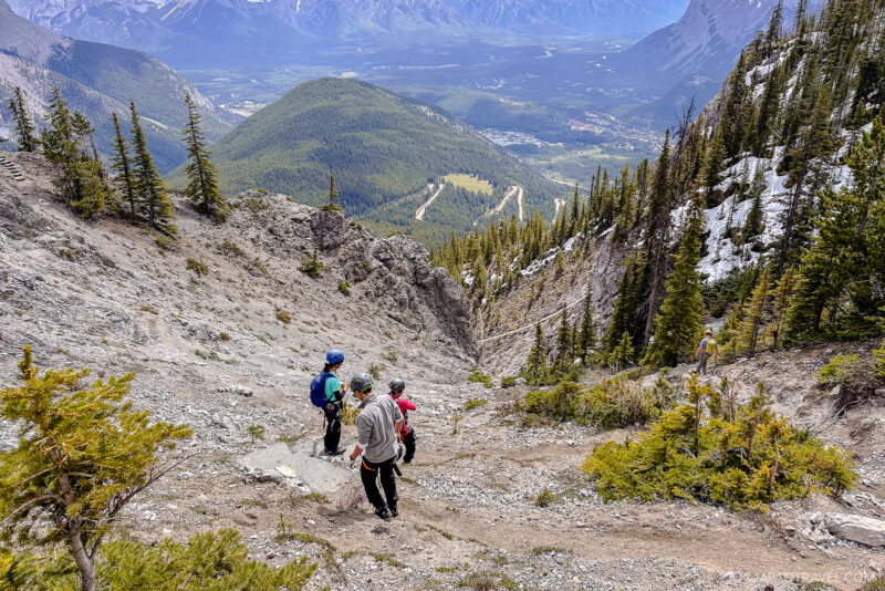 Three hikers descend a rocky mountain trail surrounded by pine trees in Banff National Park, with a scenic valley and distant mountains visible under a clear sky.