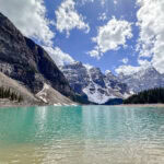 A turquoise lake in Banff National Park is surrounded by pine trees, rocky shores, and tall snow-capped mountains under a partly cloudy sky.