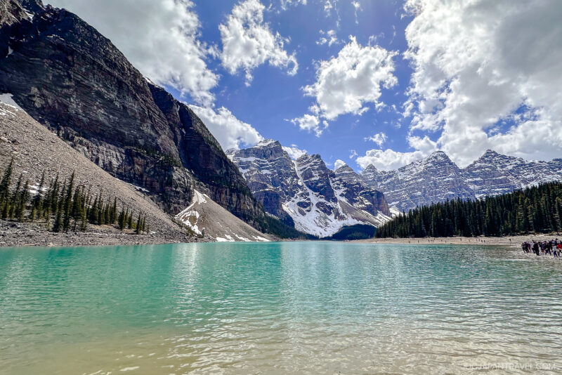 A turquoise lake in Banff National Park is surrounded by pine trees, rocky shores, and tall snow-capped mountains under a partly cloudy sky.