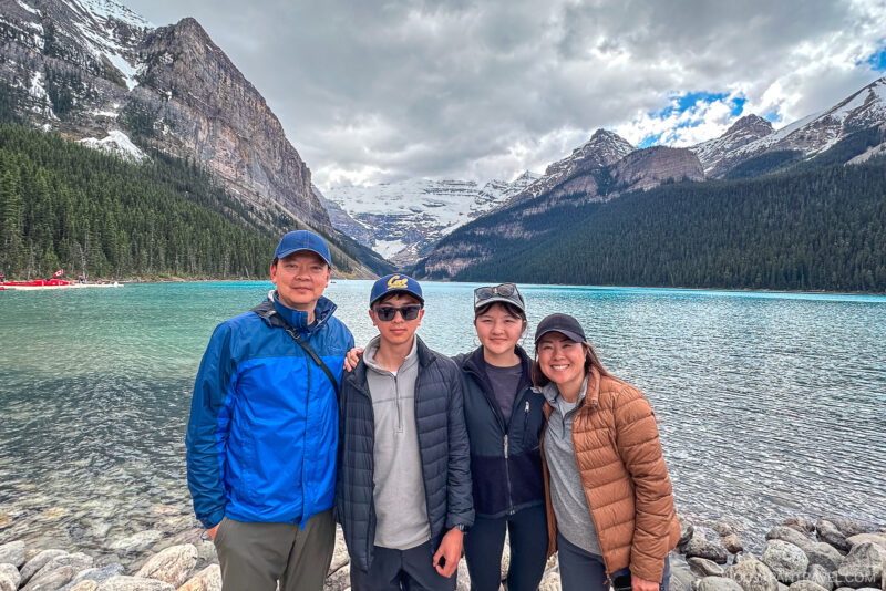 Four people pose together, smiling, in front of a turquoise lake surrounded by pine trees and snow-capped mountains under a cloudy sky in Banff National Park.