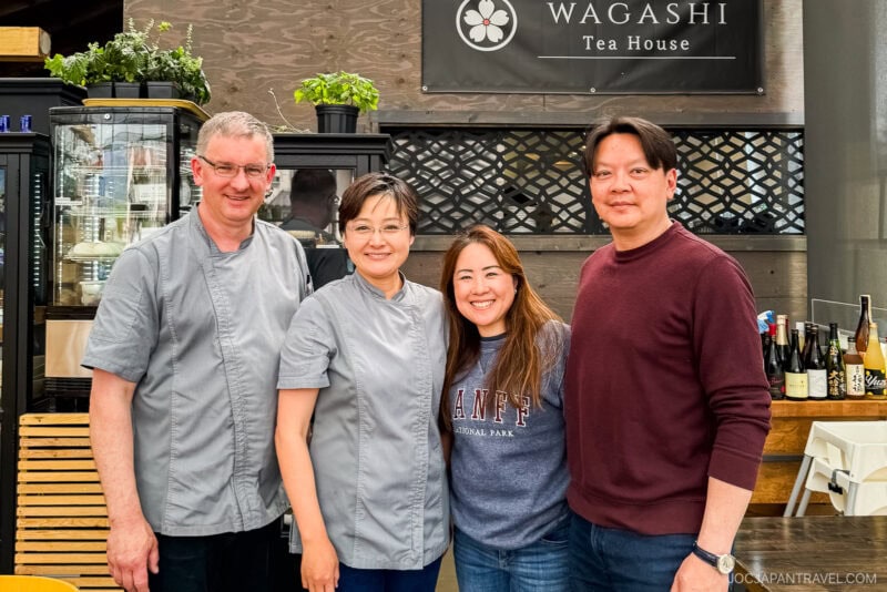 Four people are smiling and posing together inside Wagashi Tea House. Two wear grey chef jackets, while another stands in front sporting a Banff National Park sweatshirt. Bottles and shelves create a cozy background scene.