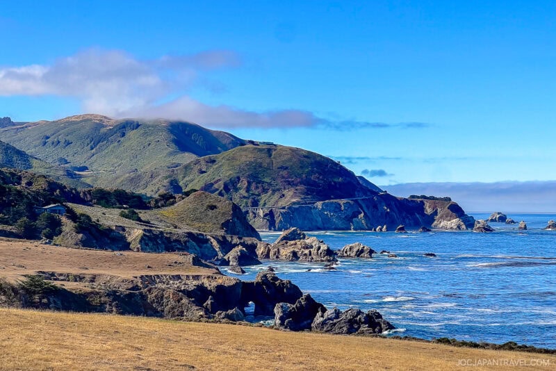 A scenic Big Sur coastal landscape features rugged cliffs, rocky shorelines, and rolling hills meeting the blue ocean under a clear sky with a few clouds drifting over the hills.