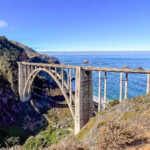 A concrete arch bridge spans a deep canyon along Big Sur’s rugged coastline, with green hills and blue ocean under a clear sky.