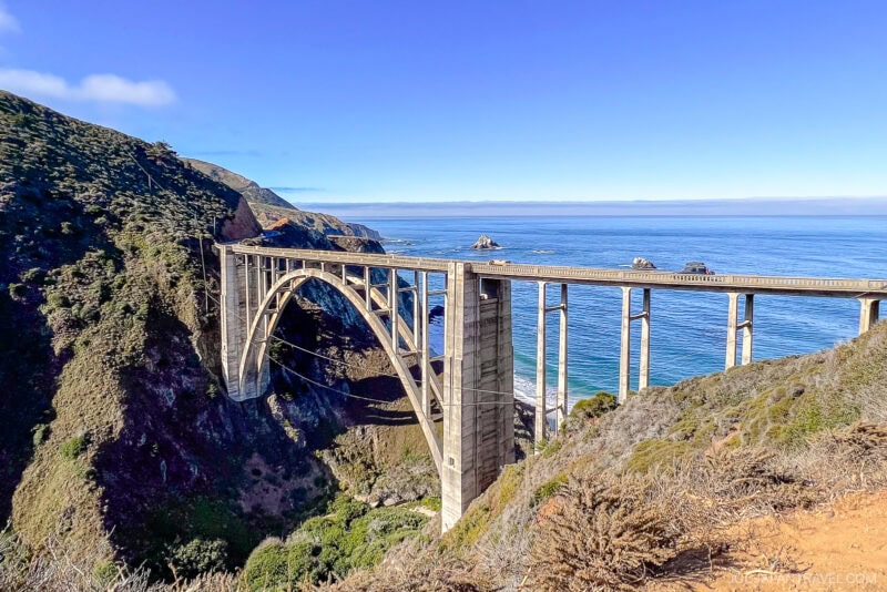 A concrete arch bridge spans a deep canyon along Big Sur&rsquo;s rugged coastline, with green hills and blue ocean under a clear sky.