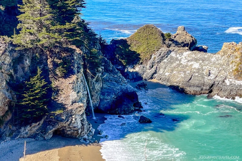 A scenic coastal view in Big Sur shows a small waterfall cascading down rocky cliffs onto a sandy cove, with turquoise ocean waves and green trees above the cliffs under a clear blue sky.