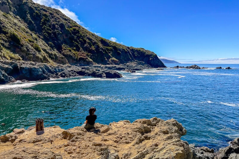 A person sits on rocky cliffs overlooking a blue ocean bay at Big Sur, with rugged hills covered in greenery rising on the left under a bright, clear sky.