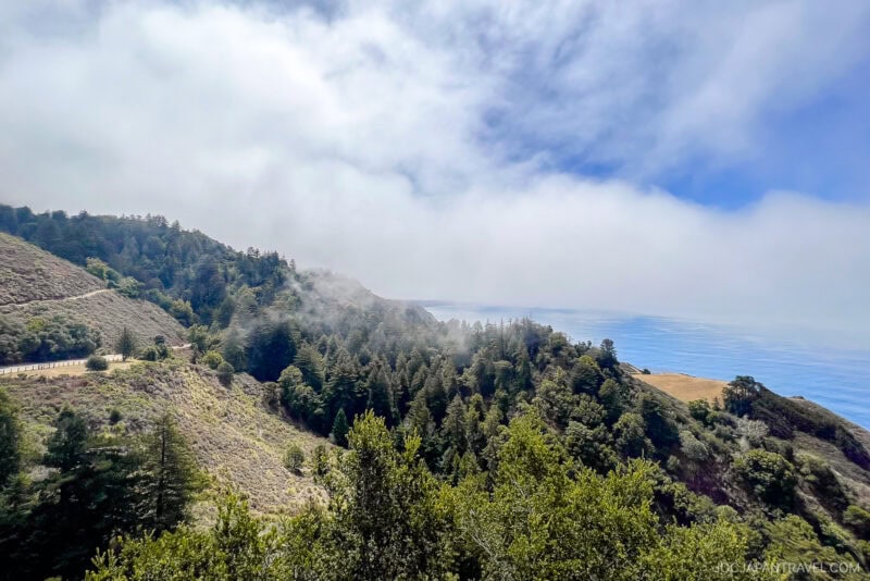 View of a coastal mountain landscape in Big Sur, with green trees, a winding road, and mist rolling in from the ocean under a partly cloudy sky.