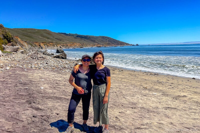 Two people stand arm-in-arm on a sandy, rocky Big Sur beach with waves and cliffs in the background under a clear blue sky.