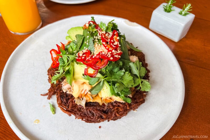 A crispy noodle pancake topped with avocado, shredded chicken, fresh herbs, sliced red chili, cucumber, and sesame seeds—served on a white plate with a glass of orange juice and a small plant, capturing the relaxed vibe of Big Sur.