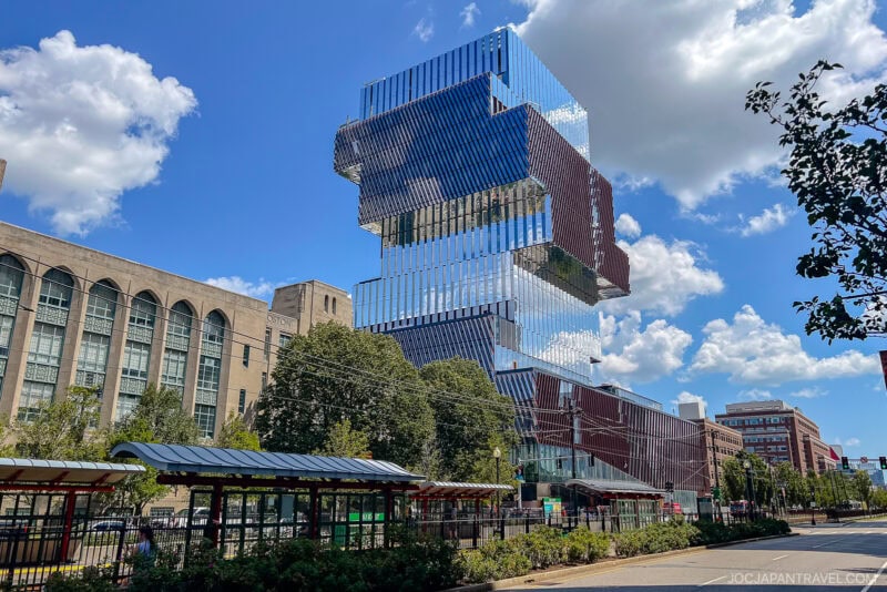 A modern, glass-covered building with stacked, offset sections rises against a blue sky with clouds—an iconic sight on the Northeast College Tour—surrounded by trees and city infrastructure, including a tram stop in the foreground.