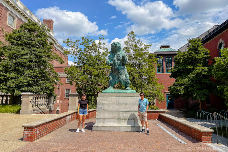 Two people stand on either side of a large bear statue on a stone pedestal in an outdoor plaza during their Northeast College Tour, surrounded by trees and brick buildings under a partly cloudy sky.