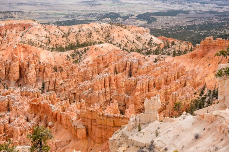 View of Bryce Canyon’s striking orange and pink hoodoos, with scattered green trees and a distant valley visible under a partly cloudy sky.